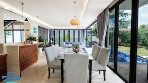 A modern dining area with a glass table, chairs, and a bottle of wine. There is a reception desk and a large window with a view of a pool and greenery.