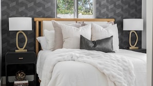 A neatly made bed with white bedding and multiple pillows, flanked by two bedside tables with lamps, in a room with patterned wallpaper and a view through large windows.