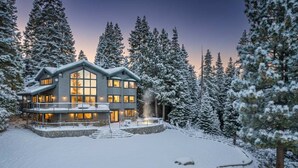 Una casa grande y moderna con múltiples ventanales y un cimiento de piedra, rodeada de árboles cubiertos de nieve.