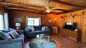 A cozy room with wooden walls and ceiling, a ceiling fan, a sofa set, a television, and a small table with a lamp.