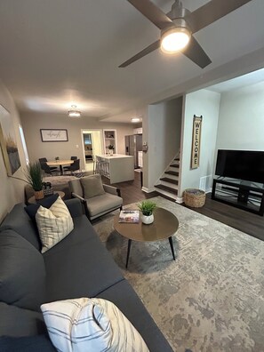 A modern living room with a gray sofa, a coffee table, and a TV. There is a staircase leading to another level, and a kitchen area is visible in the background.
