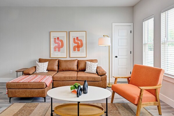 Una sala de estar con un sofá marrón, un sillón naranja, una mesa de centro blanca y dos obras de arte abstracto enmarcadas en la pared.
