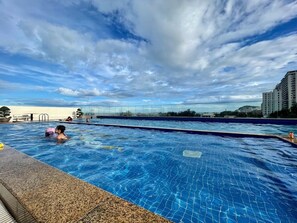 Una piscina con agua cristalina azul, rodeada por una solana con baldosas y vista a edificios al fondo.