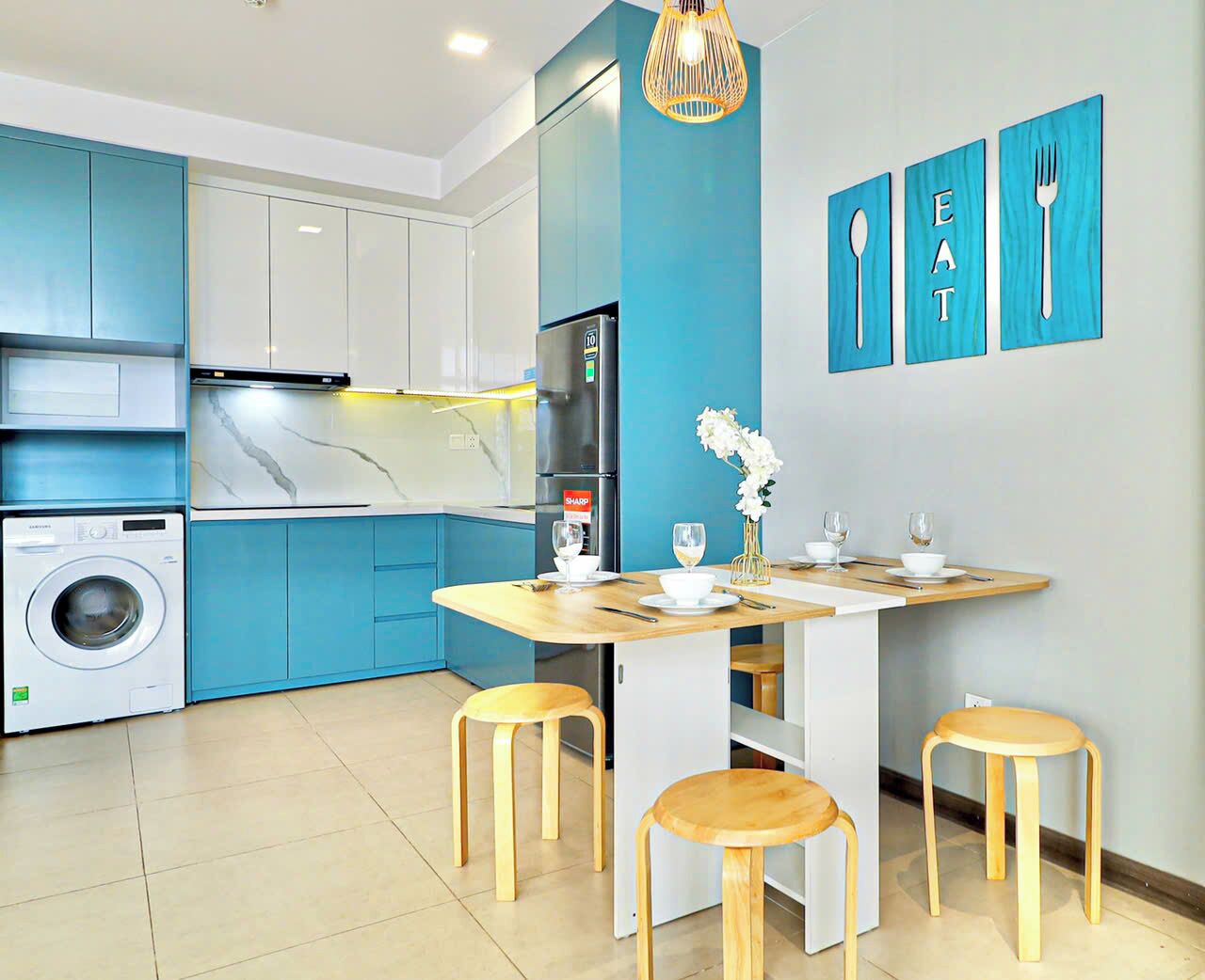 A modern kitchen with blue cabinets, a built-in washing machine, a refrigerator, and a dining area with stools and a table.