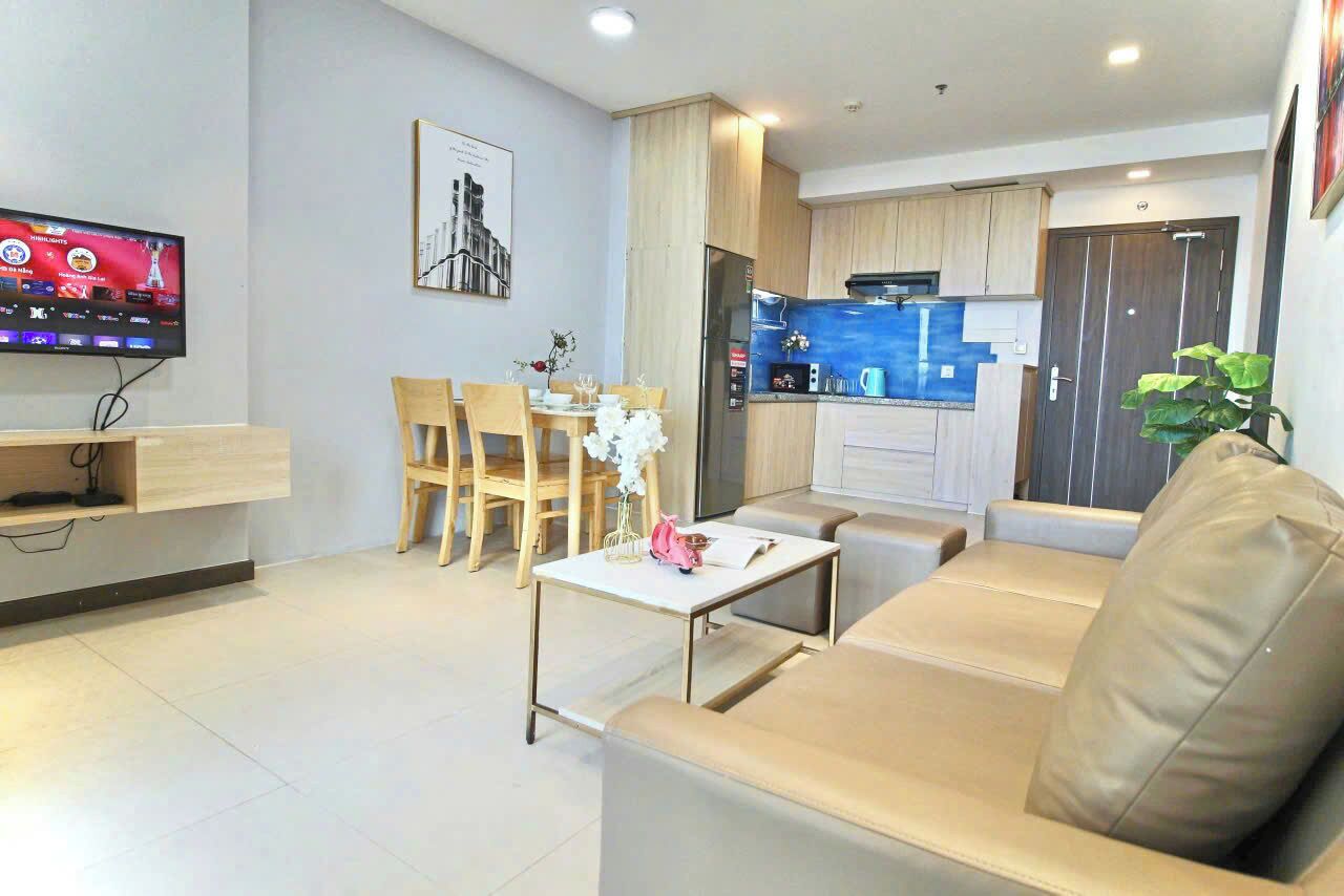 A modern living room with a sofa, a dining table with chairs, a TV mounted on the wall, and a kitchen area with cabinets and a refrigerator.
