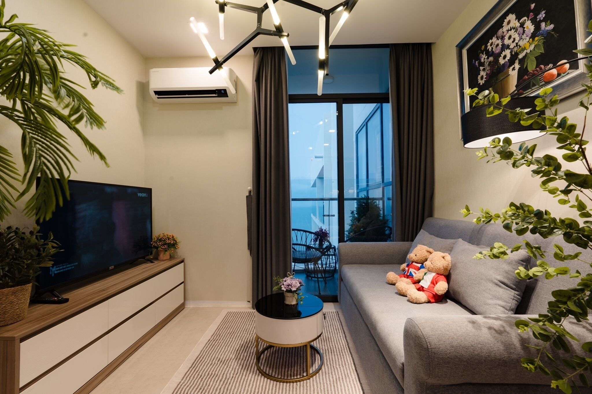 A modern living room with a flat-screen TV, a sofa with a teddy bear, a small table with a vase, and a large window with a view of the outside.
