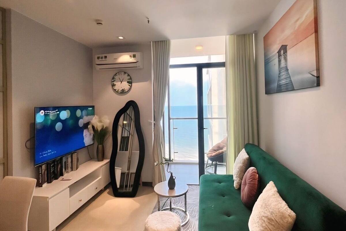 A modern living room with a green sofa, a flat-screen TV, a large mirror, and a view of the ocean through a glass door.