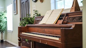 Un piano de cola con partituras en una habitación con piso de madera, un reloj en la pared y una ventana con luz natural.