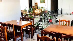 Een eetruimte met houten tafels en stoelen, een trap en uitzicht op een woonkamer met banken en planten.