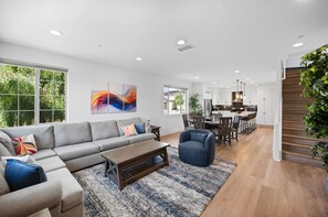 Ein modernes Wohnzimmer mit einer grauen Eckcouch, einem Couchtisch und einem blauen Sessel. Es gibt einen Essbereich mit einem Holztisch und Stühlen, und im Hintergrund eine Küche.