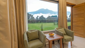A room with a large window, two green armchairs, a wooden table with fruit, and a view of a green field and mountains. - Corbett Four Seasons (Ramnagar)