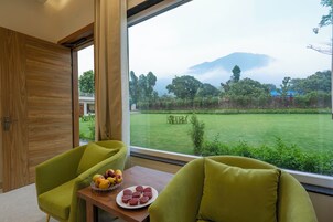 Una habitación con un ventanal grande, una puerta de madera y dos sillas verdes. Se ve una mesa con un frutero y un plato de muffins.
