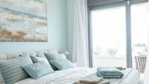A bedroom with a large bed, a ceiling fan, a view through a sliding glass door, and a beach-themed painting on the wall.