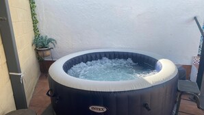 Uma banheira de hidromassagem com o logotipo da marca visível, rodeada por um terraço revestido de azulejos com uma cadeira e um vaso de planta.