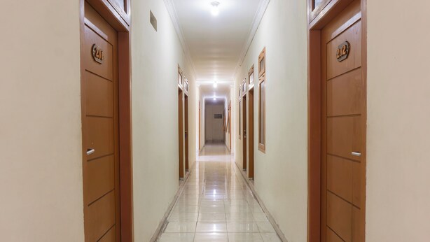 Un pasillo largo y estrecho con puertas de madera a ambos lados, cada una con un número. El suelo está cubierto de baldosas y las paredes pintadas de un color claro.