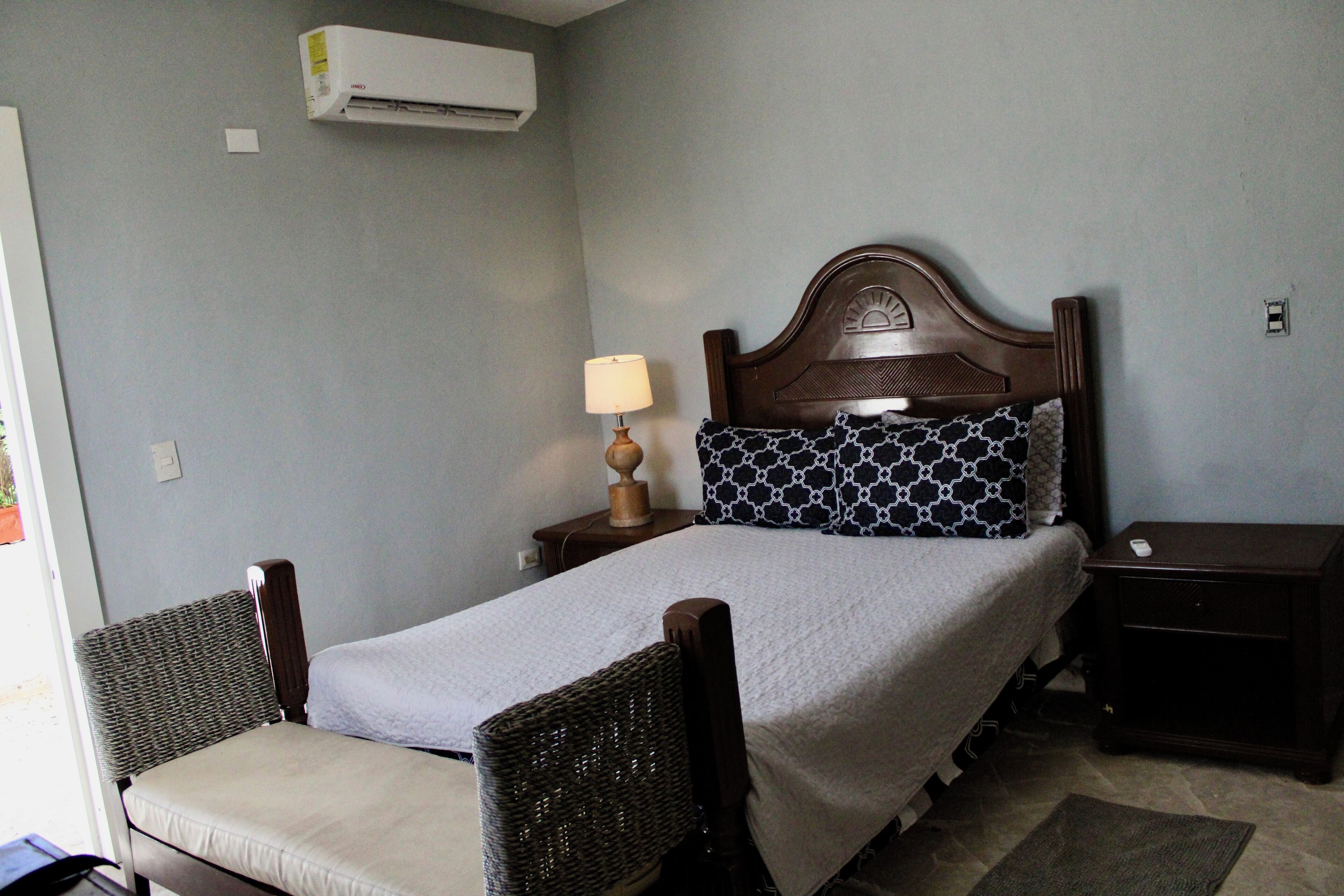 Quarto com cama, mesa de cabeceira, luminária, cadeira e ar condicionado.
