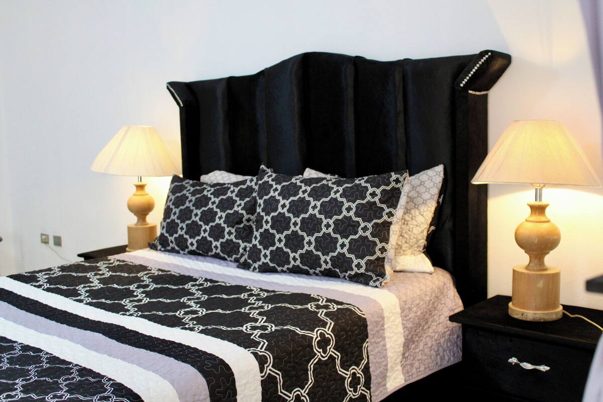 Een net opgemaakt bed met beddengoed met een patroon, twee bedlampjes en een zwart hoofdeinde.