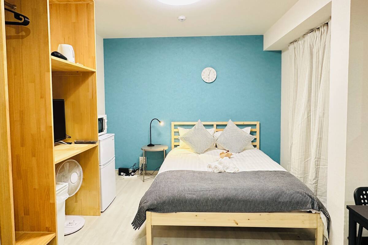 A bedroom with a bed, a wooden wardrobe, a small table with a lamp, a fan, and a clock on the wall.