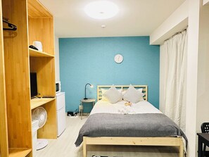 Een slaapkamer met een bed, een houten kledingkast, een tafeltje met een lamp, een ventilator en een klok aan de muur.