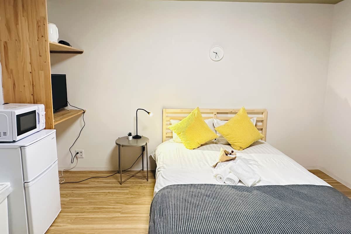 A bedroom with a bed, a bedside table with a lamp, a microwave, and a small shelf with a television.