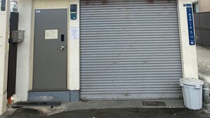Edifici amb una persiana metàllica tancada, un petit cartell a la porta i un cubell de brossa a la dreta.