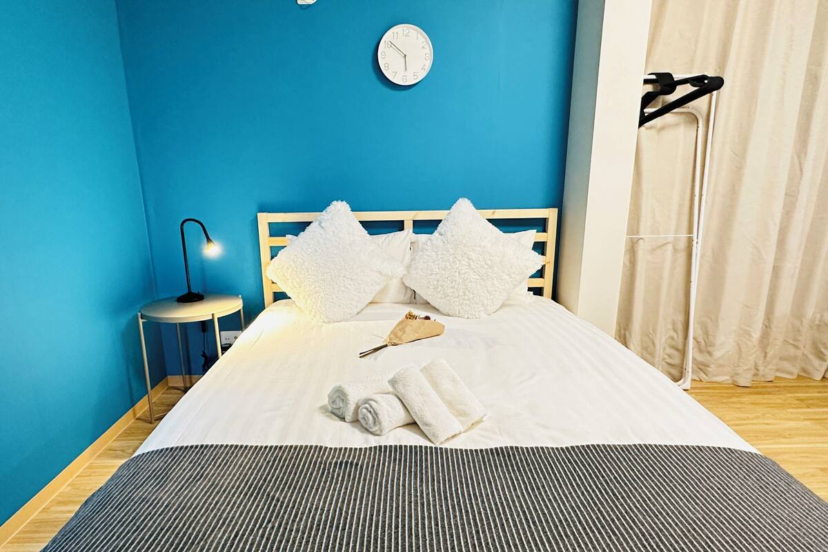 A neatly made bed with white bedding and fluffy pillows, a bedside lamp, and a clock on the wall.