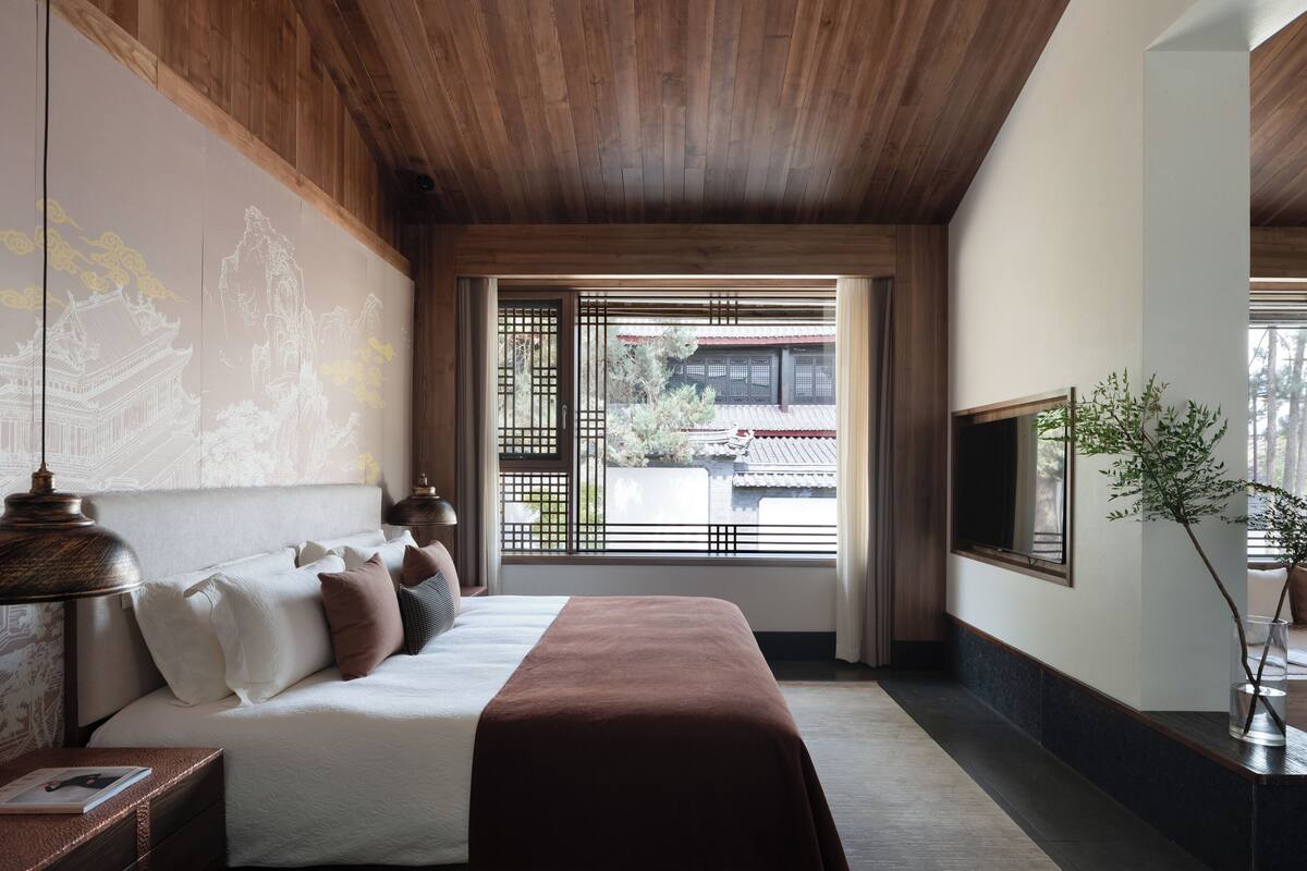 Ein Schlafzimmer mit Bett, Nachttisch, Lampe und Blick auf ein traditionelles Gebäude durch ein Fenster mit Holzgitterwerk.