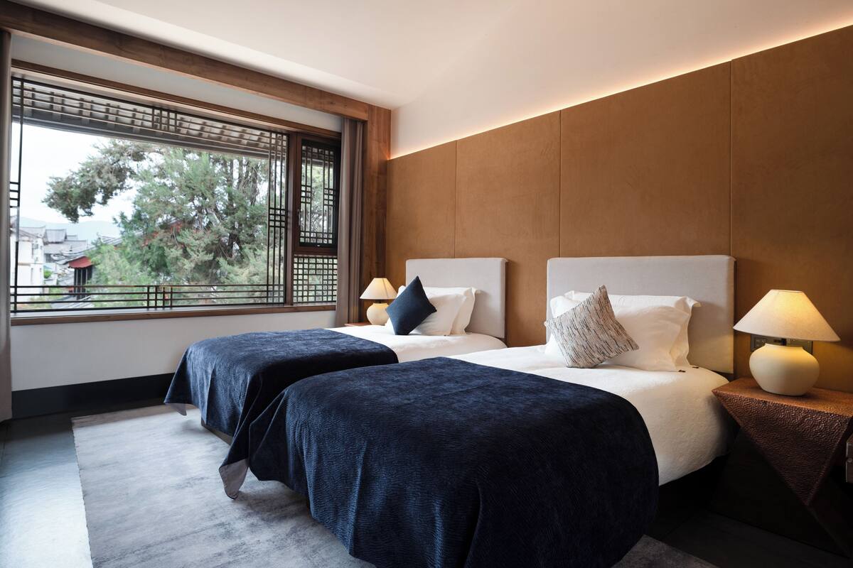 Ein Hotelzimmer mit zwei Betten, einem großen Fenster mit Blick auf Bäume und Gebäude sowie einem modernen Design mit Holzpaneelen und einem grauen Teppich.