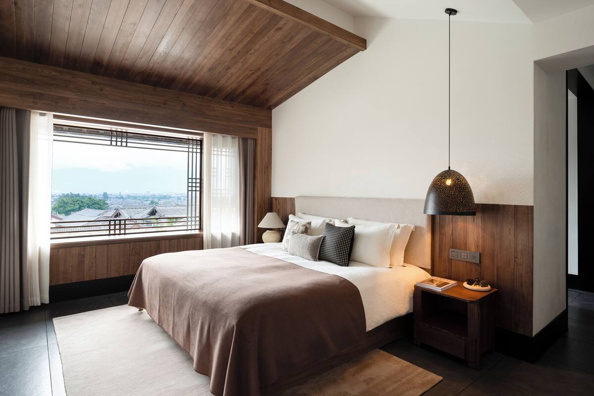 Ein modernes Schlafzimmer mit einem großen Bett, Holzboden und Blick auf eine Stadtlandschaft durch ein Fenster.