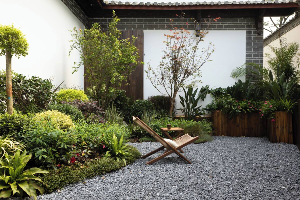 Ein Garten mit einem Holzstuhl und einem kleinen Tisch, umgeben von verschiedenen Pflanzen und einem Kiesweg.