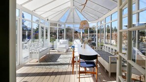 Une véranda dotée d’une terrasse en bois, de meubles blancs et de grandes fenêtres.