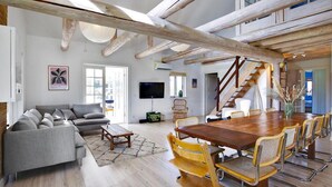 Un vaste salon avec un canapé gris, une table à manger en bois et des chaises jaunes. Un escalier mène à l’étage supérieur.