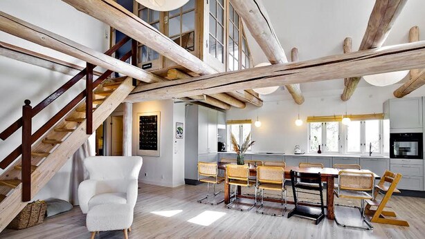 Un loft spacieux avec des poutres apparentes, un coin repas doté d’une grande table et de chaises, et un escalier menant à un niveau supérieur.