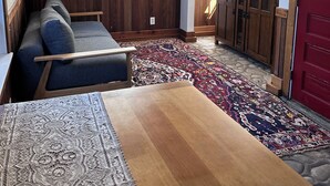 A cozy room with a wooden ceiling, a large window with a view, a sofa, a patterned rug, and a wooden table.