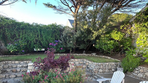 A garden with a stone border, white Adirondack chairs, and various plants and flowers.