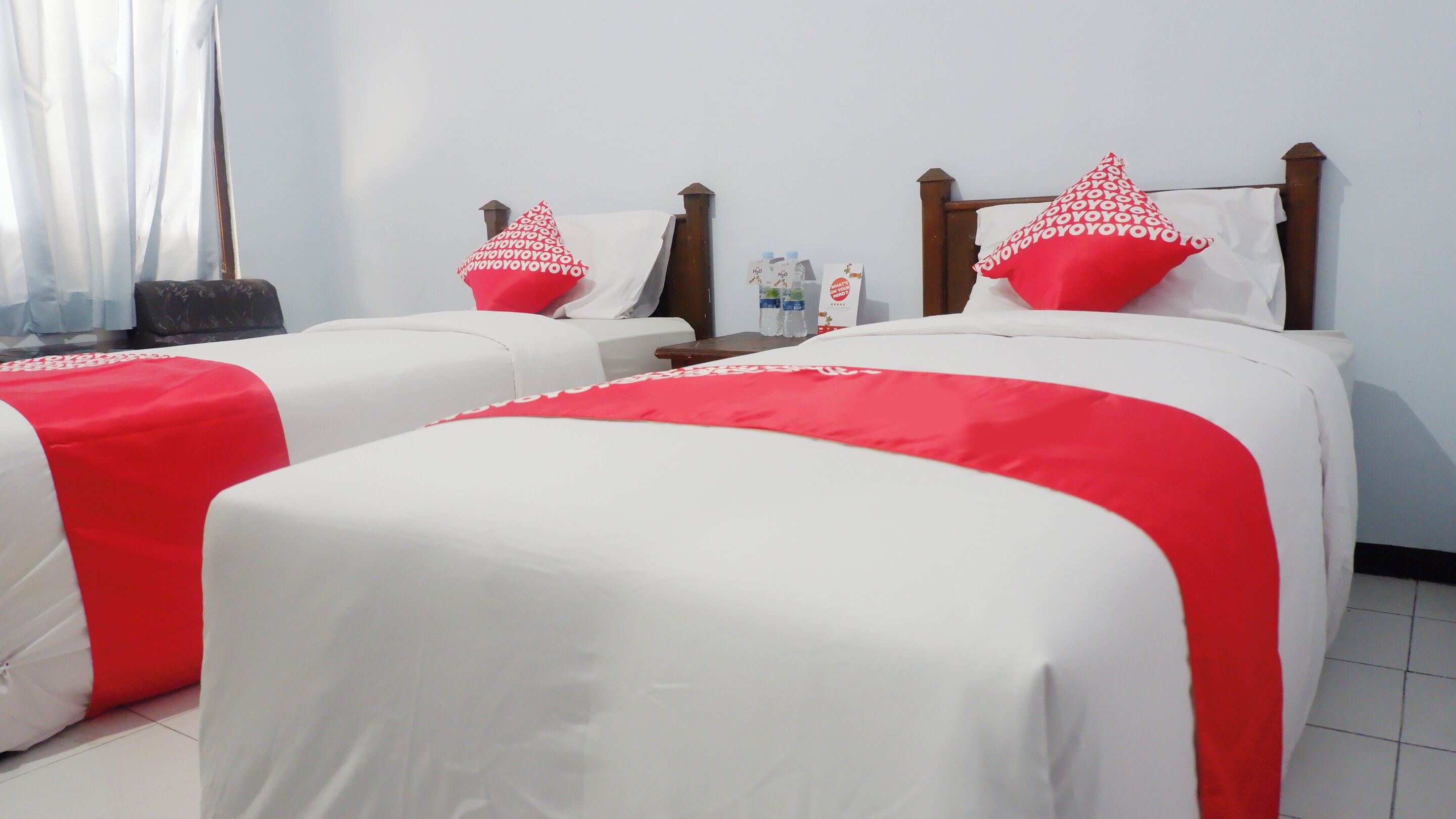 Habitación de hotel con dos camas, cada una con una colcha blanca y almohadas rojas. Se ve un mesita de noche con una lámpara y una botella de agua.