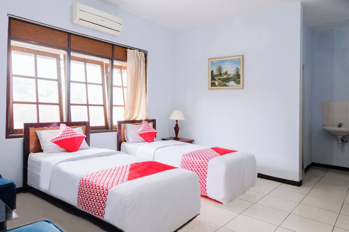 A hotel room with two beds, a window with curtains, a painting on the wall, and a sink in the corner.