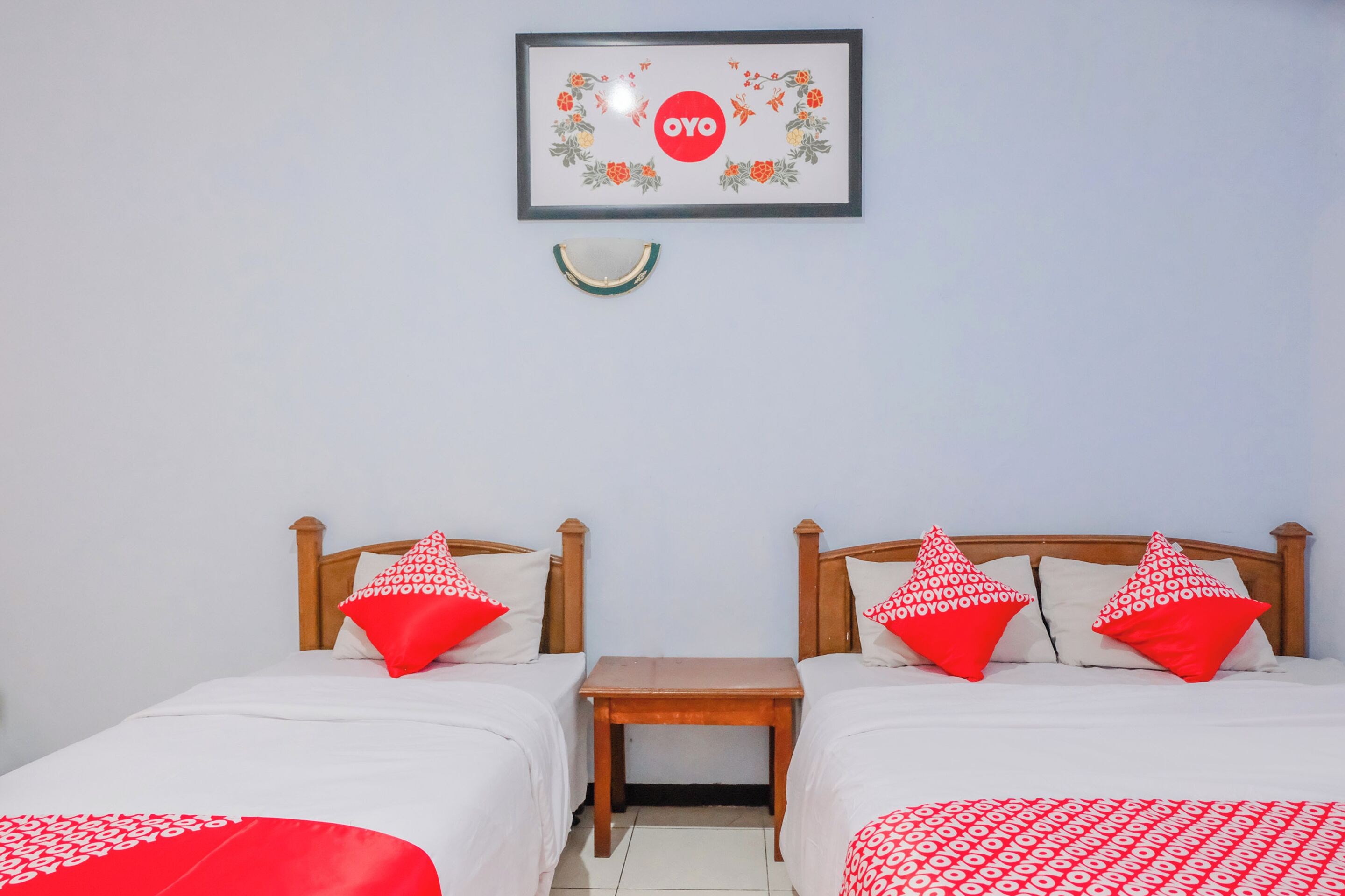 Habitación de hotel con dos camas, cada una con cabecero de madera, ropa de cama blanca y almohadas rojas con estampado. Entre las camas hay una mesita de madera, y en la pared, sobre la mesita, un cuadro con un círculo rojo y diseño floral.