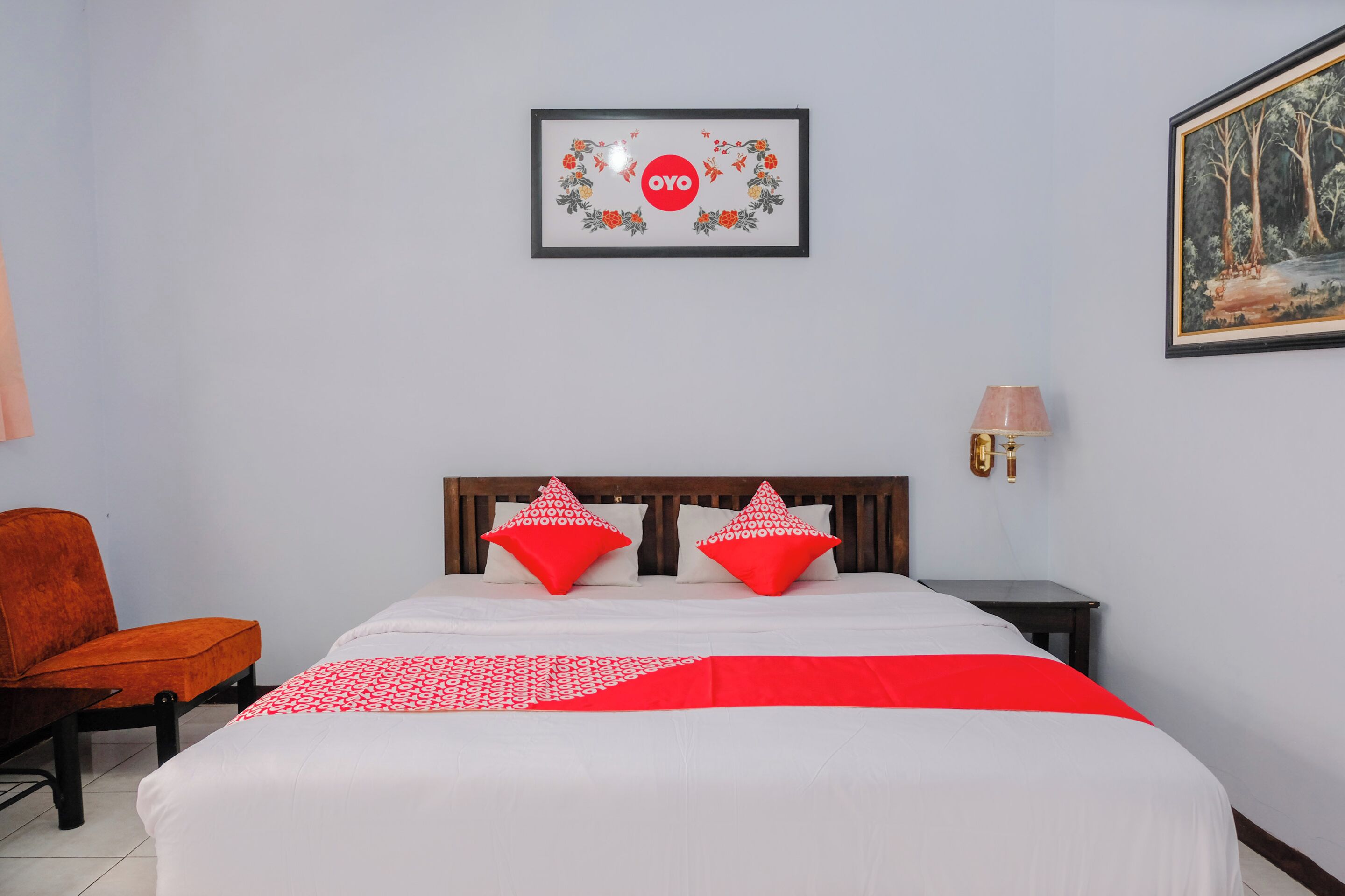 Habitación de hotel con una cama, dos almohadas rojas, un cabecero de madera, una mesita de noche con una lámpara, un cuadro enmarcado en la pared y una silla naranja.
