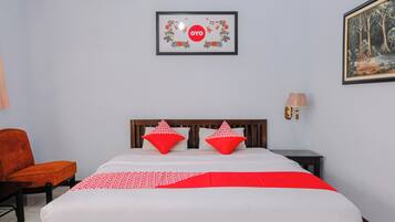 Habitación de hotel con una cama, dos almohadas rojas, un cabecero de madera, una mesita de noche con una lámpara, un cuadro enmarcado en la pared y una silla naranja.