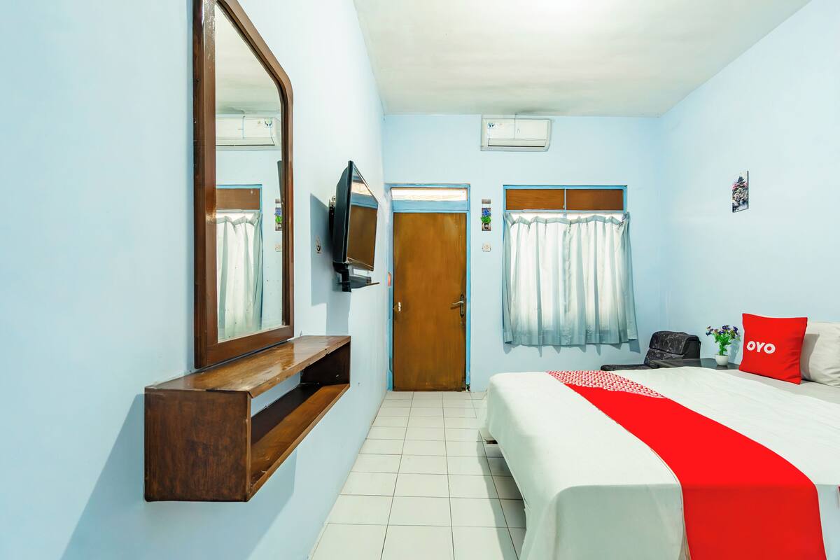 A bedroom with a bed, a TV, a wooden shelf, a door, and a window with curtains.
