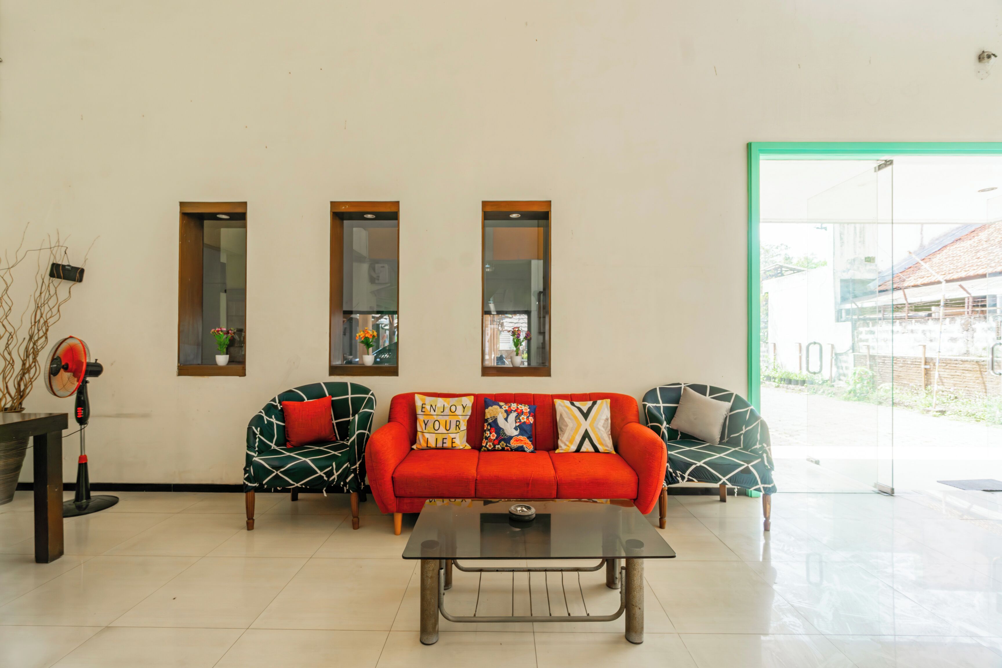 A living room with a red sofa, two patterned armchairs, a glass coffee table, and three windows with blinds.