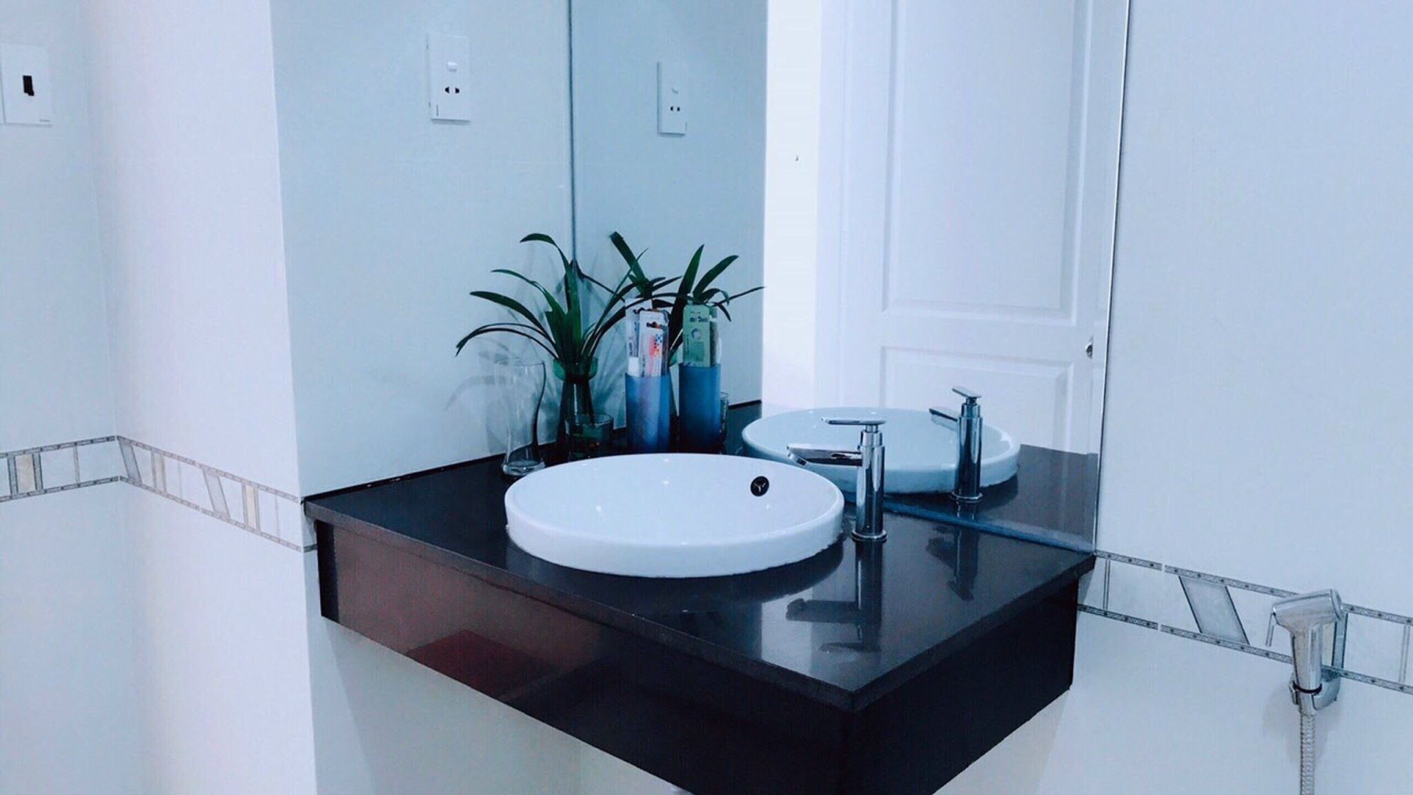 A bathroom with a white sink, a mirror, and a black countertop. There are two plants on the counter and a glass of water.