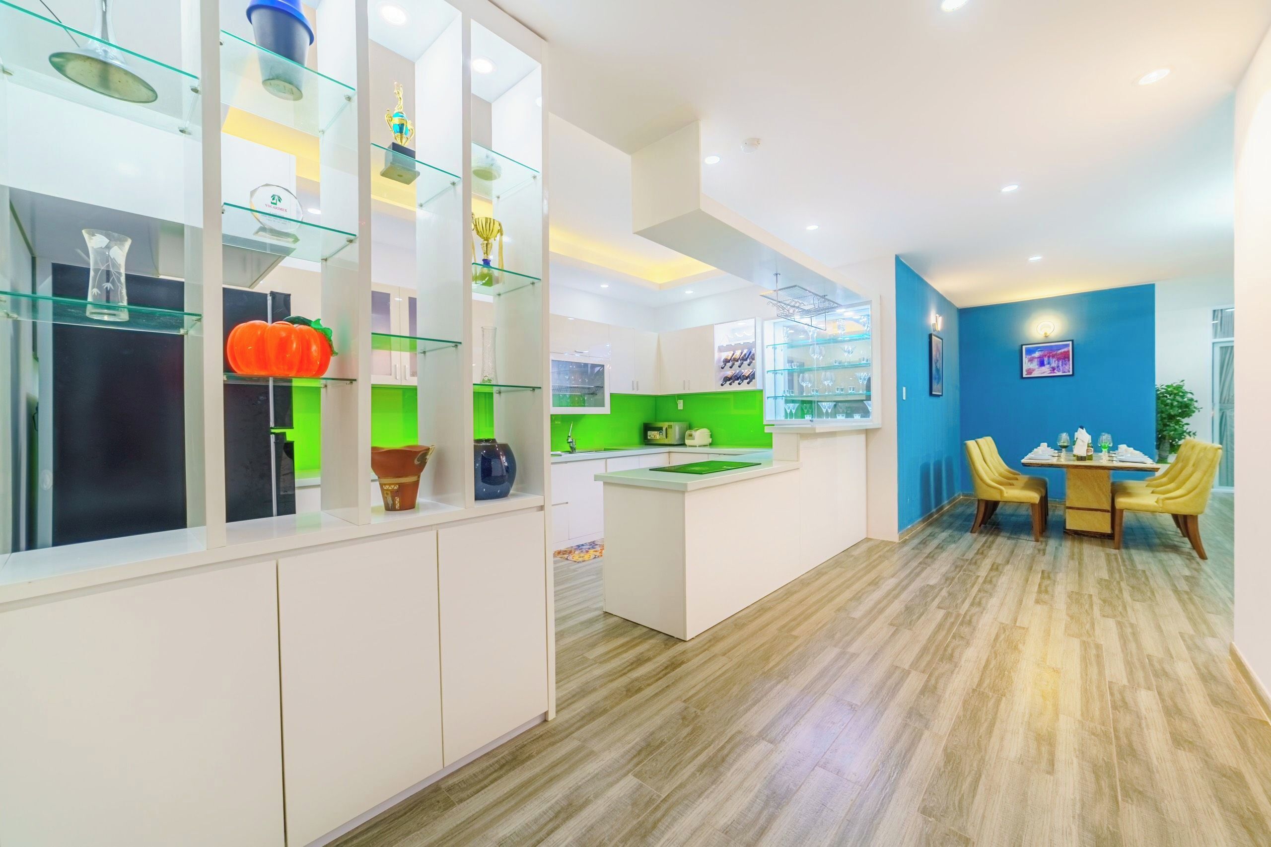 A modern kitchen with glass shelves, a central island, and a dining area with yellow chairs.