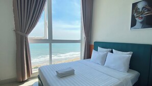 Une chambre d’hôtel dotée d’une grande fenêtre donnant sur l’océan, d’un lit parfaitement fait avec du linge de maison blanc et d’une tête de lit de couleur turquoise.