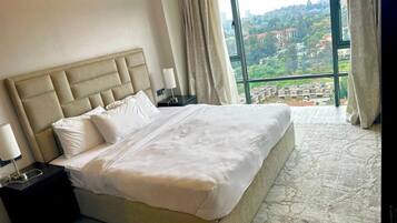 A hotel room with a large bed, a nightstand, a lamp, and a view of a cityscape through a large window.