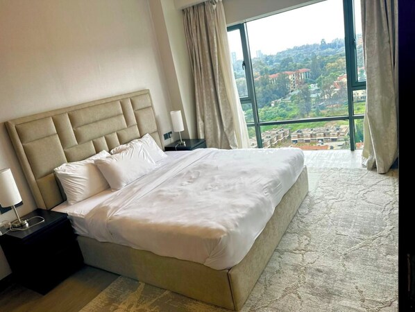 A hotel room with a large bed, a nightstand, a lamp, and a view of a cityscape through a large window. - Escada Apartments (Nairobi)