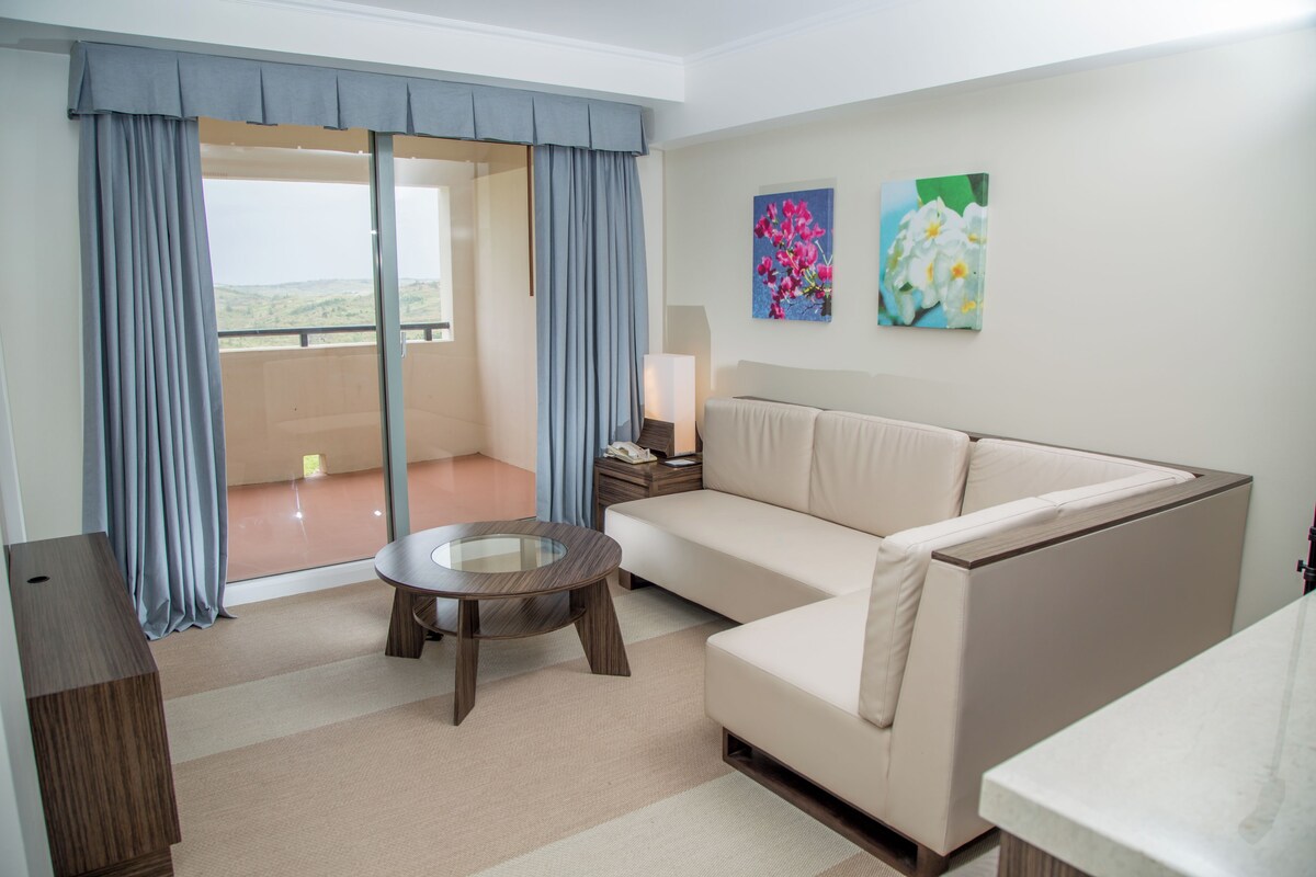A modern living room with a sofa, a glass-top coffee table, and a view of the outdoors through large sliding doors.