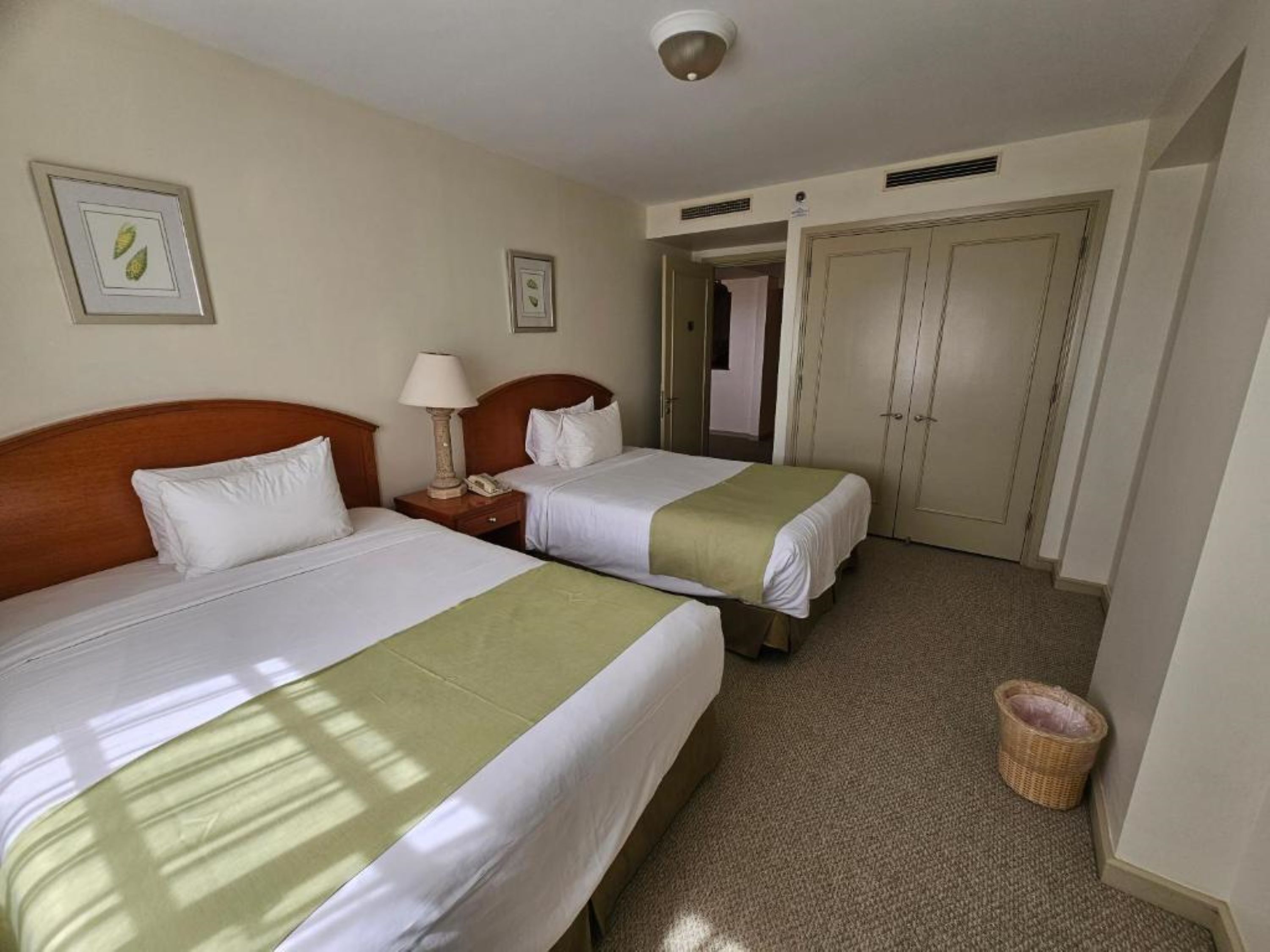 A hotel room with two beds, a nightstand, a lamp, a telephone, a waste basket, and a door leading to another room.