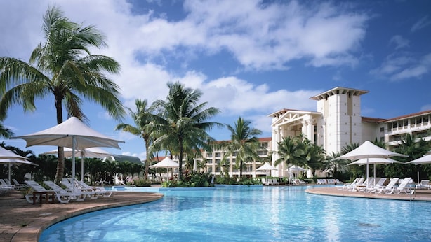 A resort pool area with lounge chairs and umbrellas, surrounded by palm trees and a hotel building in the background.